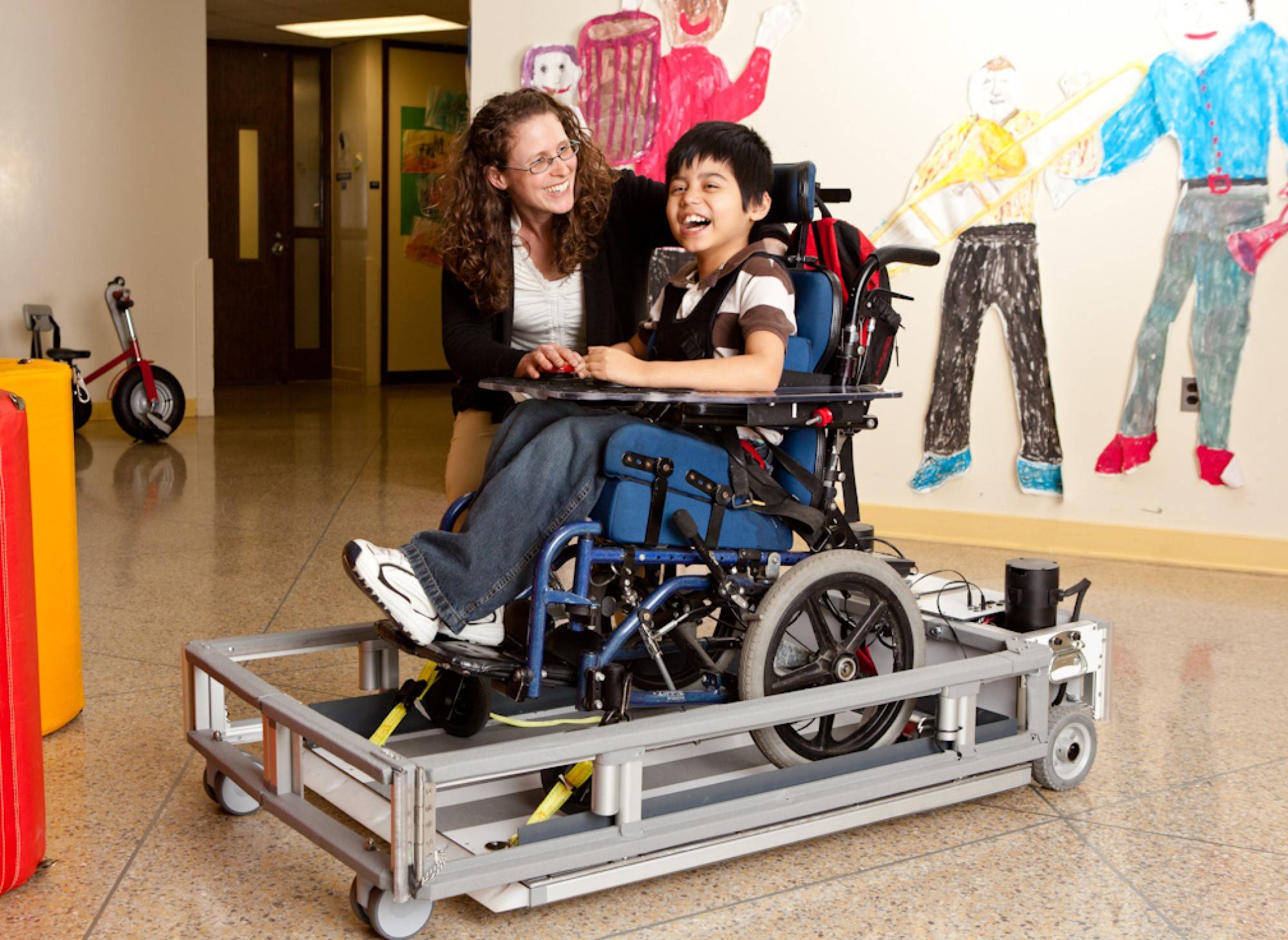 a woman kneels next to a child in a mobility aid